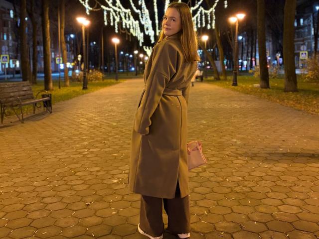JeseniaPiontek with brown coat, black tights, white shoes, pink purse, standing on cobbled path, looking back, city lights, tree lights in background, night, her ass slightly visible.