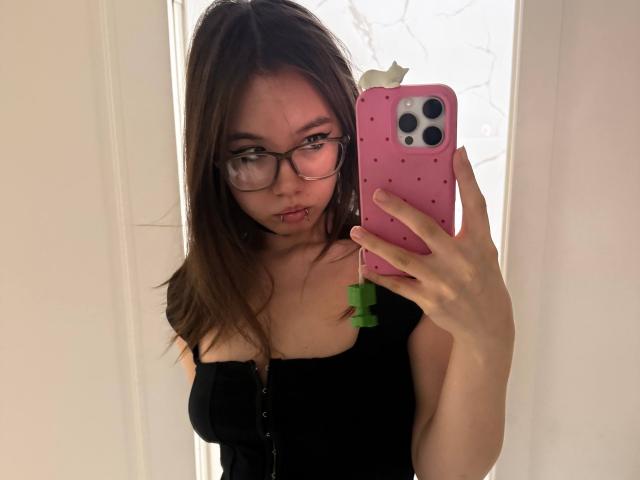 EmelineLattig with glasses, brown hair, pouty lips, black top, holding pink phone with green clip, looking in mirror. Boobs visible under top. Reflective white background.