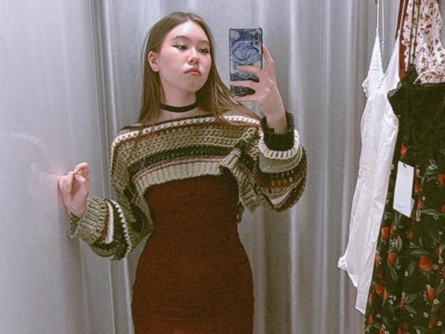 EmelineLattig with long brown hair, wearing a striped green and brown sweater, brown dress, black choker, holding phone in mirror, standing in dress store, showing her tits slightly.