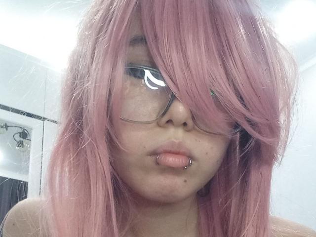 EmelineLattig with pink hair, glasses, and a lip piercing, showing her pouty, slightly chubby face and small, unadorned tits, with a neutral, slightly dirty expression.