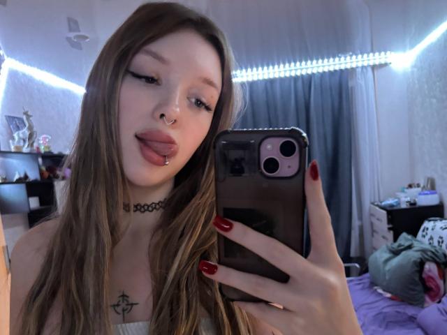 RossanaSemone with long brown hair, nose ring, and tongue out, holding a phone with red nails, showing a chest tattoo, wearing a choker, in a bedroom.