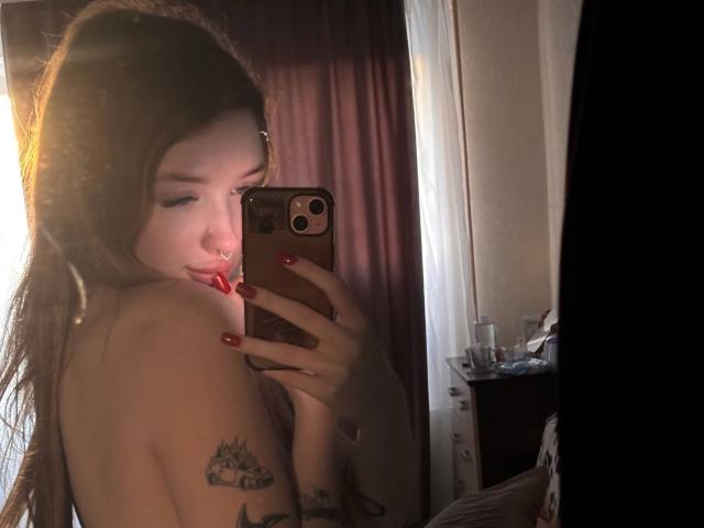 RossanaSemone with long brown hair, nose ring, red nails, and tattoos, taking a mirror selfie, showing her bare shoulder, holding a phone with a gold case.