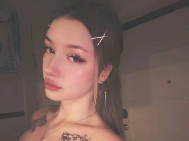 RossanaSemone with long brown hair, gold cross hairpins, hoop earrings, nose ring, tattoos on chest, makeup, pink lips, looking at camera, dimly lit bathroom.