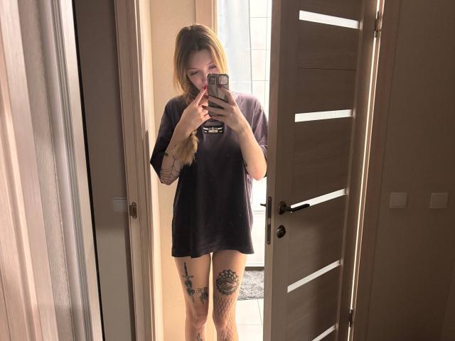 RossanaSemone with long blonde hair, wearing a black t-shirt, holding a phone, showing tattoos on her left thigh and right calf, standing in a mirror selfie.