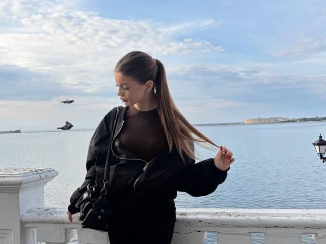 BethanySandhoff with long brown ponytail, wearing black sheer top, black jacket, and black bag, standing by white railing, holding hair, blue sky, and two birds in background.