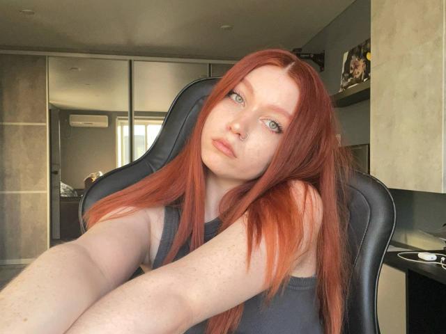 LilliamChevalier with long red hair, pale skin, and freckles, wearing a black tank top, sitting in a black chair, taking a selfie with a neutral expression.