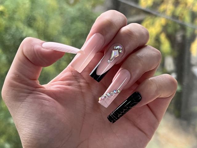 Valnes' hand with long, pink, and black acrylic nails, featuring a silver heart and diamond accent on the ring finger, against a blurry outdoor background.