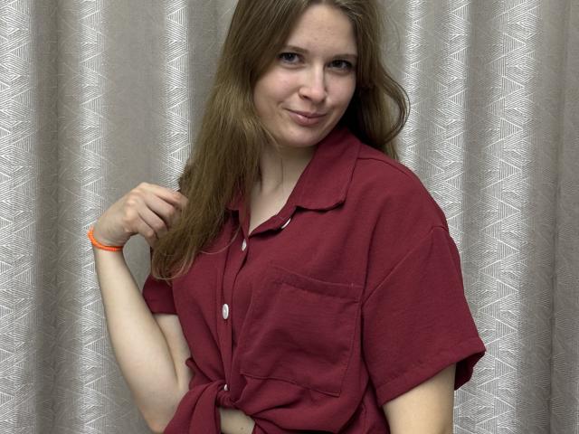 GussiePlatania, com cabelo castanho claro, vestindo uma camisa vermelha de botões, sorrindo levemente, com a mão direita perto do cabelo, pulseira laranja no pulso esquerdo, olhando para a câmera..