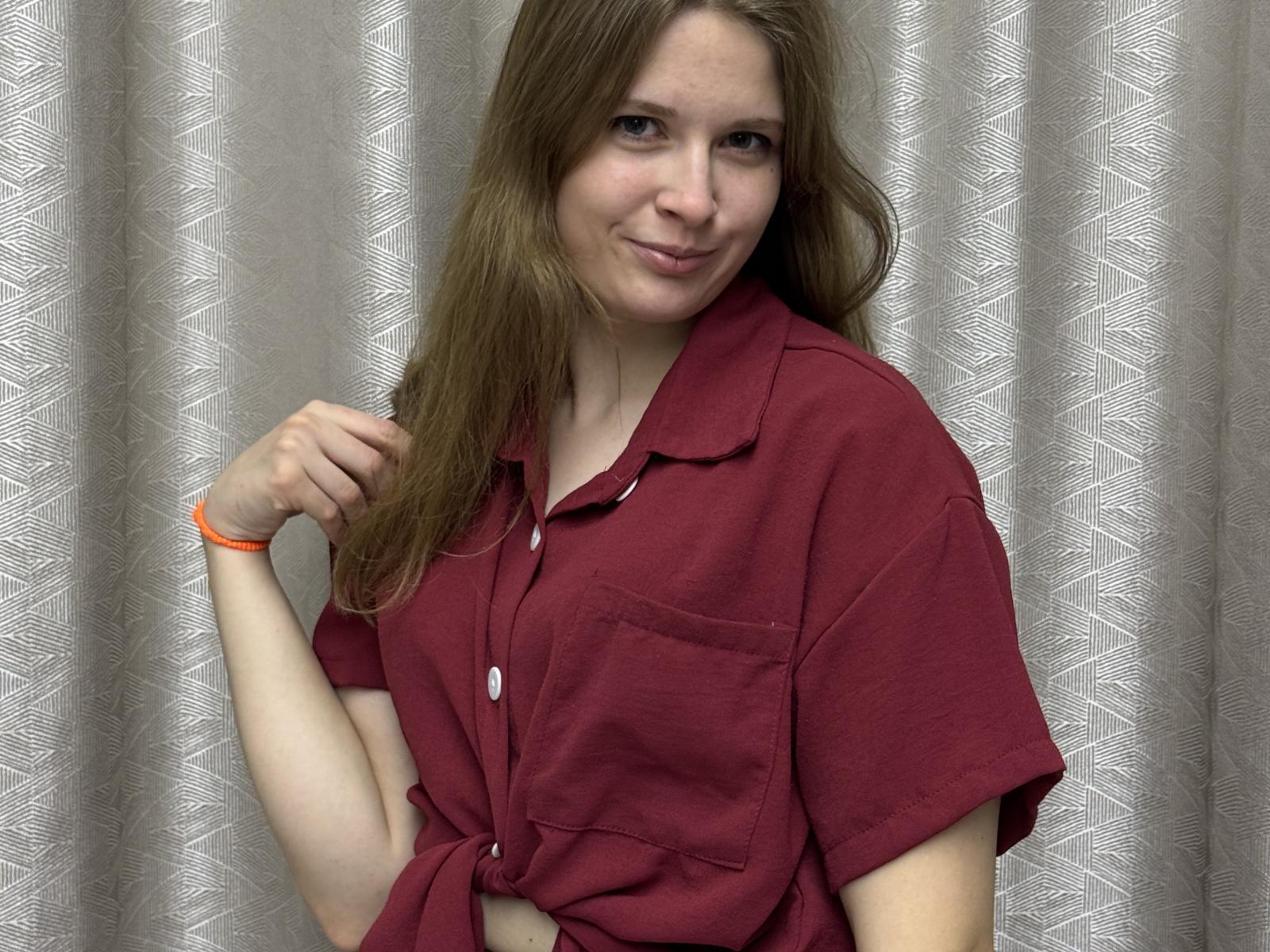 GussiePlatania, con i capelli castano chiaro, indossa una camicia rossa con bottoni, sorride leggermente, la mano destra è vicino ai capelli, ha un braccialetto arancione al polso sinistro, e guarda verso la fotocamera..