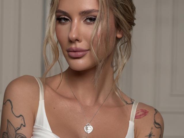 SensualMila, con i capelli biondi, tatuaggi sulle spalle, indossa una maglietta bianca e una collana d'argento con un ciondolo a forma di cuore, e guarda la fotocamera con un sorriso appena accennato..