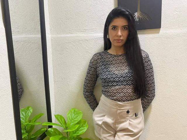 MilusSky with long black hair, wearing a lace top showing her small tits and beige satin pants, standing against a white wall, hands in pockets, looking at camera.
