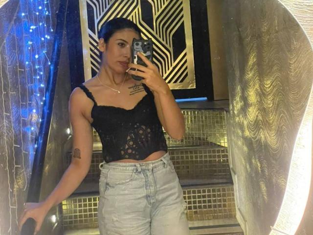 MilusSky with dark hair, black lace top, and light jeans, holding phone, tattoo on left arm, blue string lights on left, gold patterned wall in background.