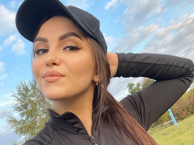 AnettLo with pouty lips, wearing a black cap and long-sleeve, has her brown hair in a ponytail, hand behind head, and brown eyebrows, blue sky in background.