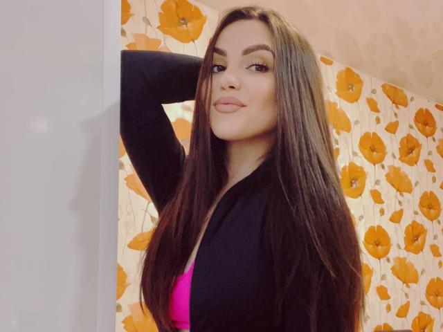 AnettLo, avec de longs cheveux bruns, un cardigan noir et un soutien-gorge rose, bras levé, regardant la caméra, avec un sourire en coin, dans une pièce avec du papier peint à motifs de fleurs jaunes..