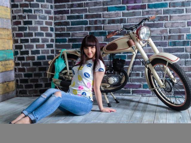 AnettLo, avec ses cheveux brun foncé, portant une chemise à motif de baiser et un jean déchiré, est assise pieds nus sur un plancher en bois, à côté d'une moto vintage beige avec un phare rond..