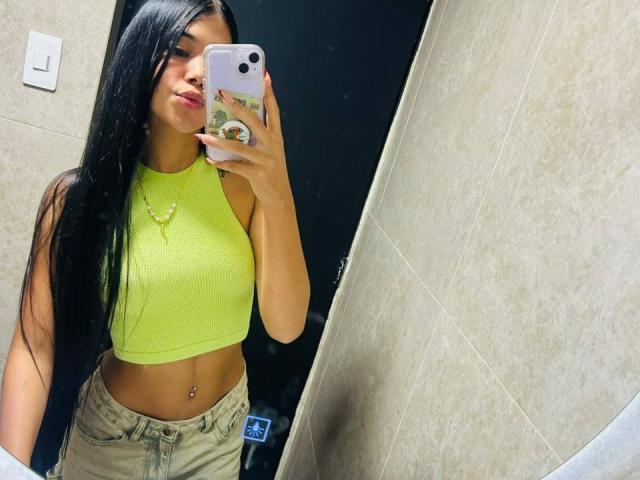 NataliaVelmonte with long black hair, neon yellow crop top, and beige high-waisted jeans, holding a phone in a mirror selfie, showing her small tits and belly button.