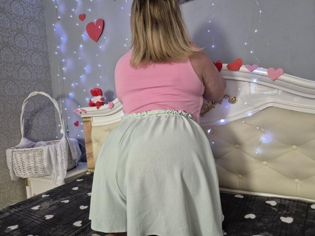 RadaRori with blonde hair, pink top, and green skirt, bending over a bed, showing her big ass, under white string lights with red heart decorations.