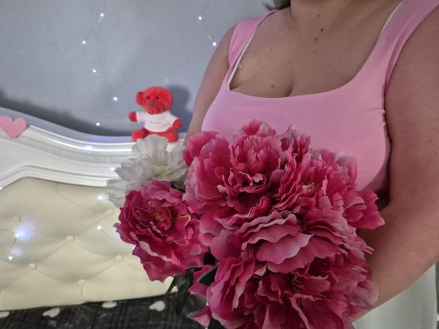 RadaRori with pink tank top, holding big pink and purple peony bouquet, her boobs visible, white tufted headboard with fairy lights, red teddy bear with heart, in background.