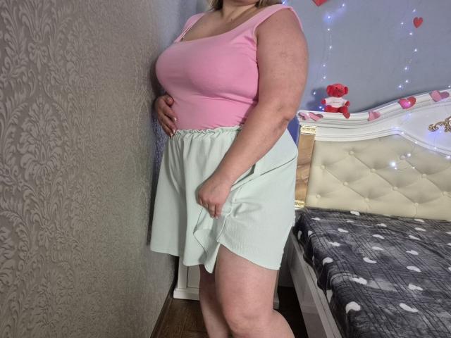 RadaRori with big tits, pink tank top, and light green shorts, standing by a bed, hand on hip, in a room with patterned wallpaper and string lights.