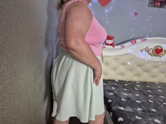 RadaRori, with a pink tank top and white skirt, stands showing her ass and a bit of her pussy, in a bedroom with a tufted white headboard.