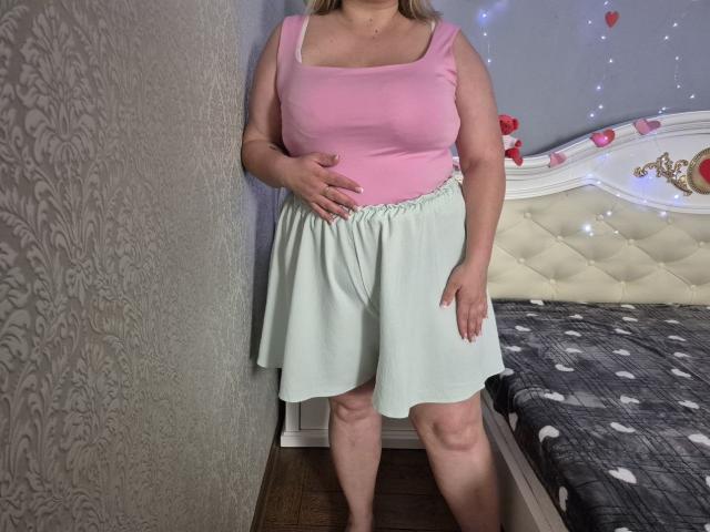 RadaRori has big boobs, a pink tank top, and a mint green skirt, standing in a room with a patterned wall and a bed with a polka dot blanket.