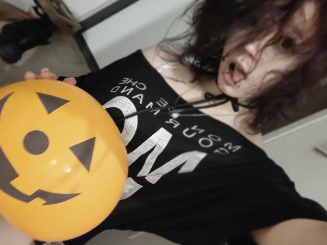 SophiaRenlay with messy brown hair, black choker, black off-shoulder shirt, holding a yellow jack-o'-lantern balloon over her small tits, mouth open, showing a slightly mischievous expression.