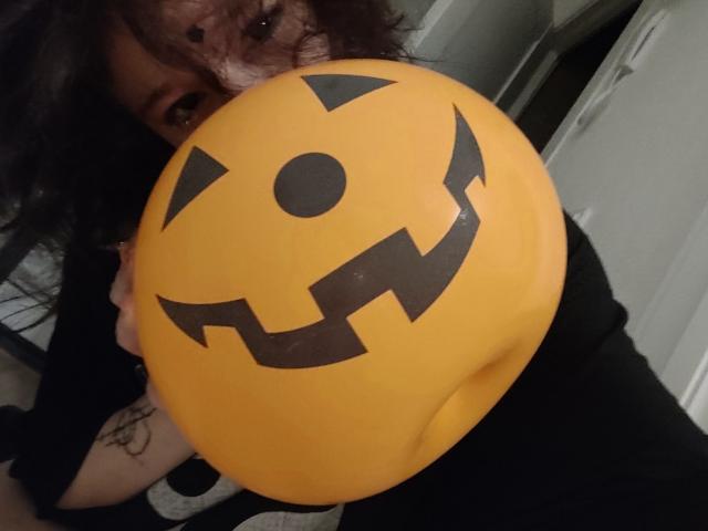 SophiaRenlay holding a yellow jack-o'-lantern balloon, her curly dark hair partially covering her face, with a black grin on the balloon. Her tattoo is visible on her arm.