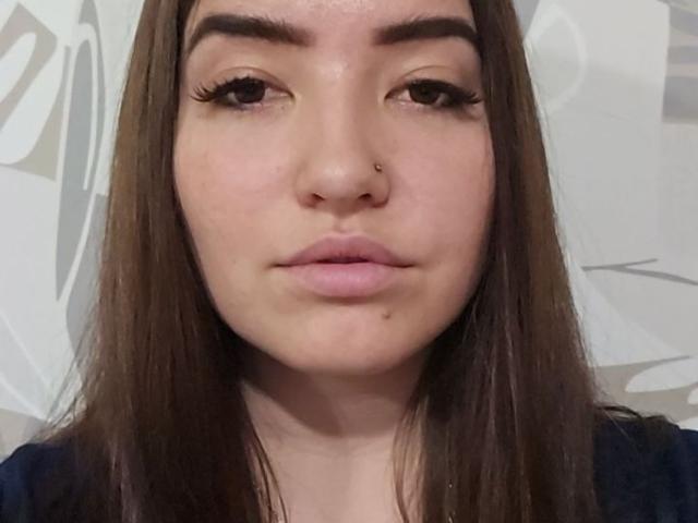 Le visage d'Emeline Minacucci, avec ses longs cheveux bruns, ses sourcils épais, son eye-liner noir et son petit piercing au nez, affiche une expression neutre, et une petite tache de naissance sous sa lèvre droite..