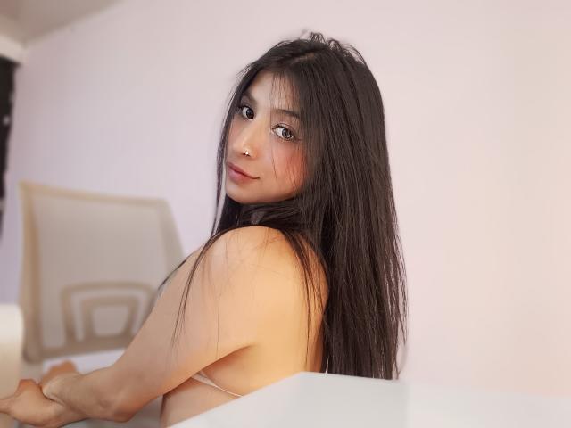 NaiaSanders with long black hair, looking back with a slight smile, showing her bare back and side-boob, sitting on a white bed. She has a nose ring.