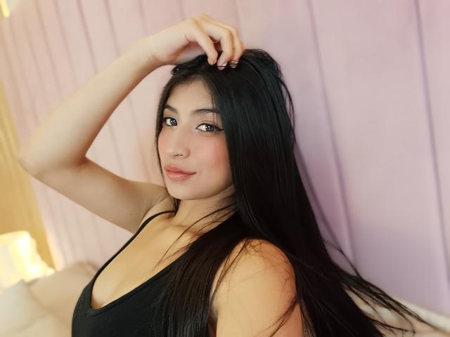 Naia Sanders, com cabelos longos e pretos, blusa de alça preta, sorriso discreto, mão no cabelo, pele clara, olhos castanhos, bochechas rosadas, iluminação suave, fofa, sexy, seios médios..