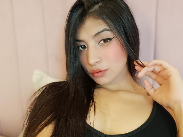 Naia Sanders, com cabelos pretos longos, pele clara, olhos castanhos, blush rosa, top preto, piercing no nariz e lábios entreabertos, segurando o cabelo, mostrando seios pequenos e um rosto fofo..