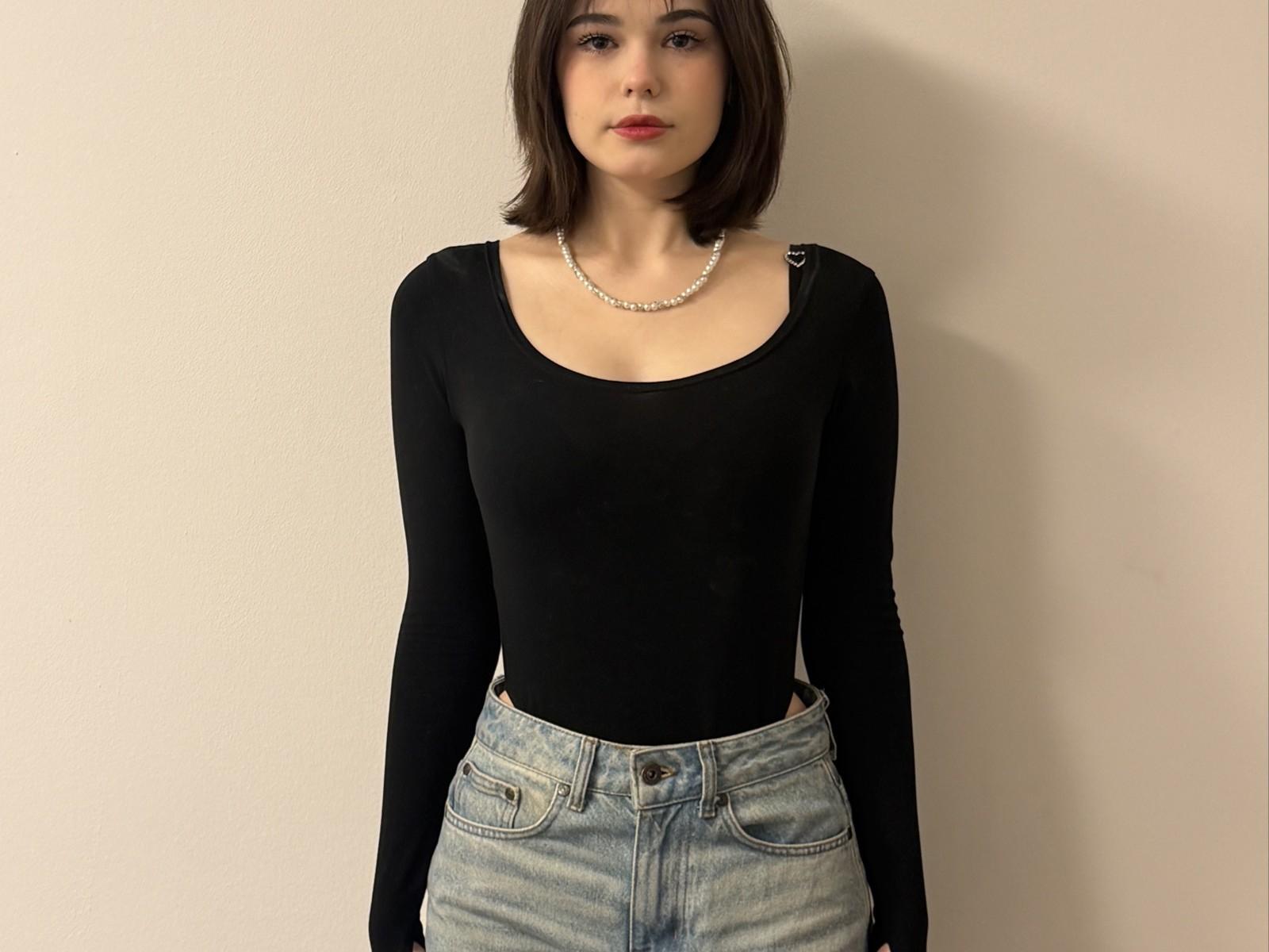 ArleenJeannoel with short brown hair, wearing a black long-sleeve top, white pearl necklace, and light blue jeans, standing against a plain wall. Boobs slightly visible under top.