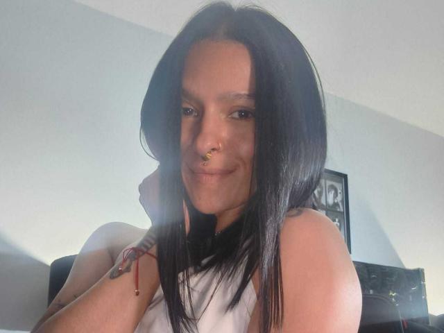 Amelie Blake, con capelli neri, piercing al naso e una canottiera bianca, sorride alla fotocamera, con la testa leggermente inclinata, un braccio appoggiato sulla sedia, un braccialetto al polso, in una luce interna brillante..