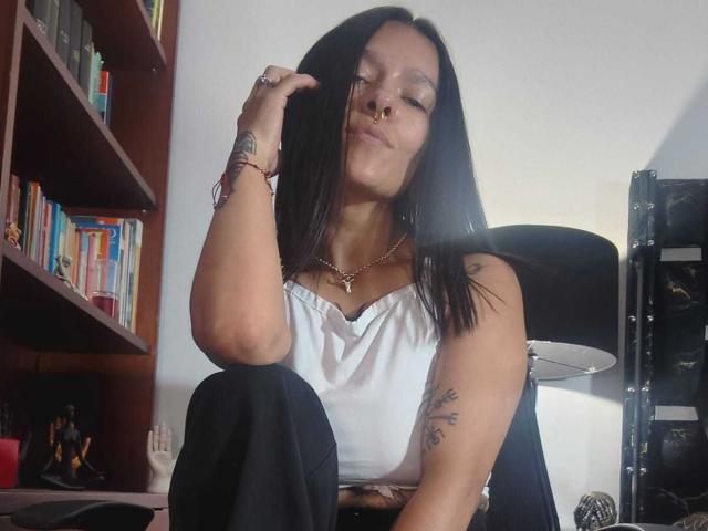 Amelie Blake, con capelli scuri, piercing al naso e una maglietta bianca, seduta a gambe incrociate, con un sorriso sardonico, la mano vicino alla testa, pantaloni neri, una libreria, una lampada e uno scaffale con libri sullo sfondo..