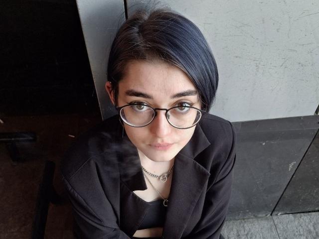 Ligia Gottshall, con i capelli neri corti, gli occhiali e una giacca nera, guarda verso l'alto, indossa una piccola collana e ha un sorriso appena accennato, con il respiro visibile in una stanza poco illuminata..