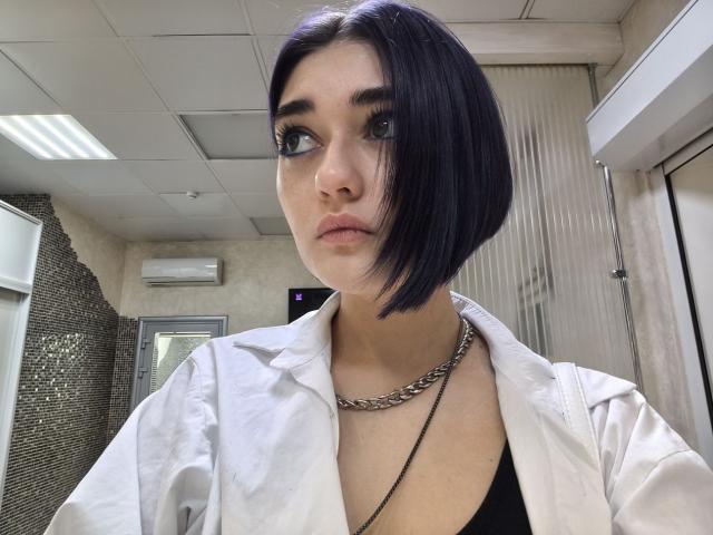 LigiaGottshall has a short black bob, wearing a white shirt over a black top, chain necklace, and heavy eyeliner, looking straight at the camera with a neutral expression.