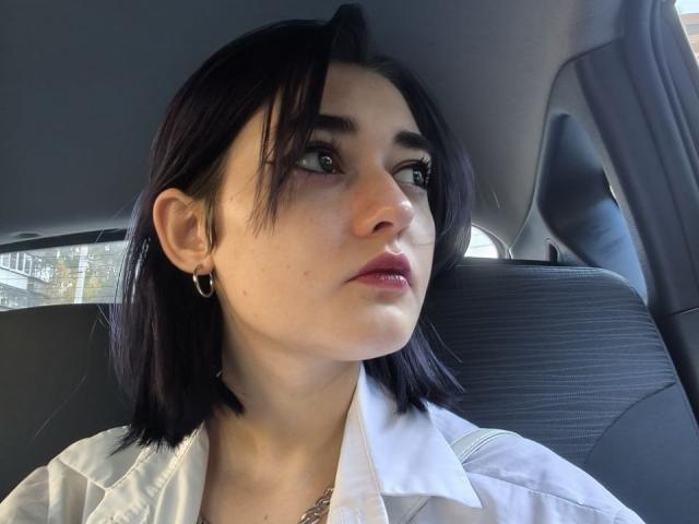 Ligia Gottshall, met donker zwart haar, kleine oorringen, een wit shirt en een gouden ketting, kijkt opzij met een neutrale uitdrukking in het grijze interieur van een auto..