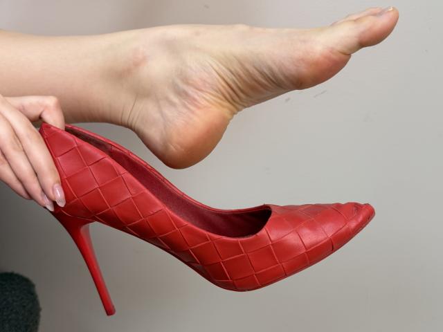 Tattiya's foot in the air, holding a red, diamond-patterned high heel with a stiletto heel, her fingers gently gripping the shoe. Her toes are slightly curled.