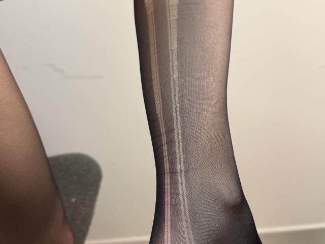 Tattiya's leg in sheer black pantyhose, showing a slight tear and visible skin underneath. Her foot is pointed downward, with a grey carpeted floor partially visible.