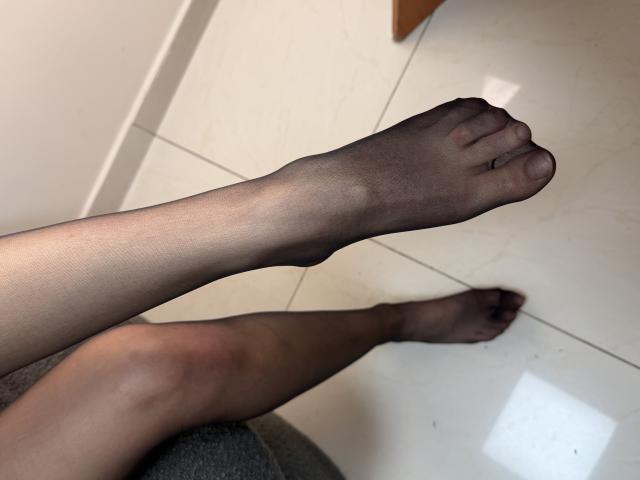 Tattiya's legs wearing sheer black stockings, with one foot raised, showing her toes. Her skin is light brown, and she's sitting on a grey surface.