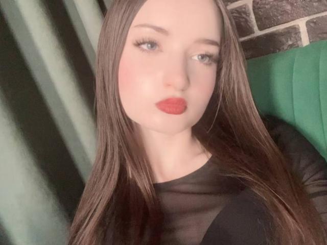 LatoshaGuarin has long brown hair, red lipstick, and light makeup, wearing a black sheer top, with blue eyes and a serious expression. Her tits are not visible.