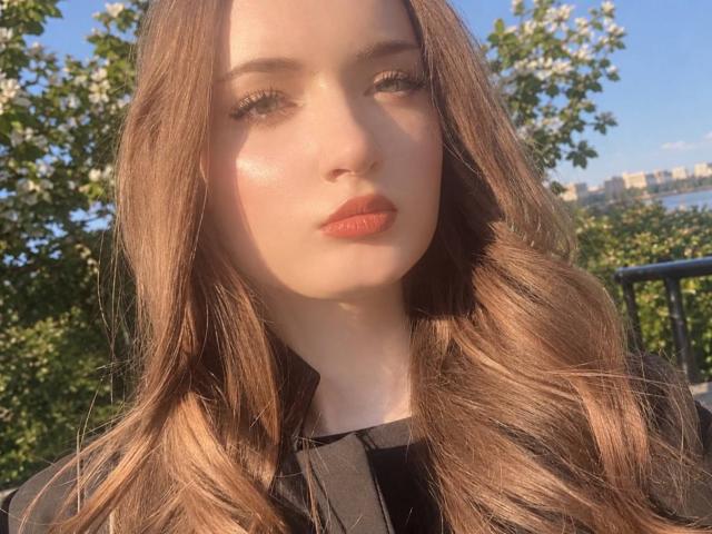 LatoshaGuarin with wavy brown hair, fair skin, red lipstick, and black top, looking serious in sunlight, greenery in background, blue sky. Boobs not visible.