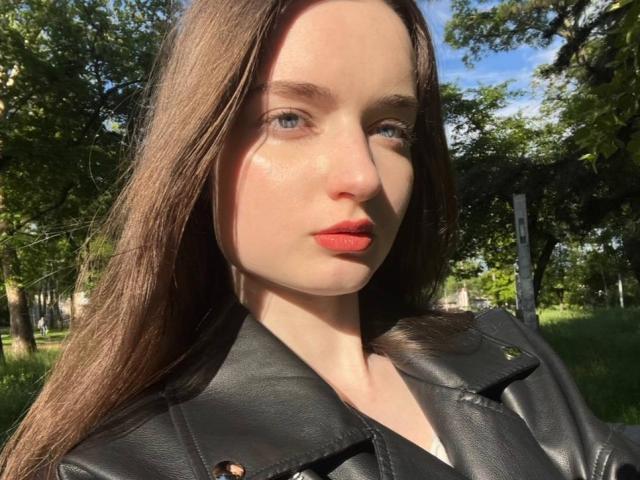 LatoshaGuarin has pale skin, long brown hair, blue eyes, and bright red lips. She's wearing a black leather jacket, sunlight highlighting her face. She's outdoors, surrounded by trees.