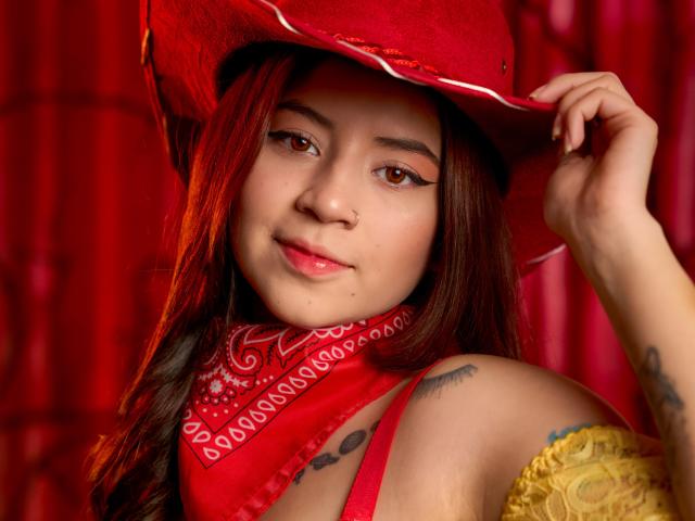 EmmaFeri with long brown hair, red cowboy hat, red bandana, yellow off-shoulder top, tattoos on shoulder, brown eyes, pink lips, holding hat, red background, cute, sexy.