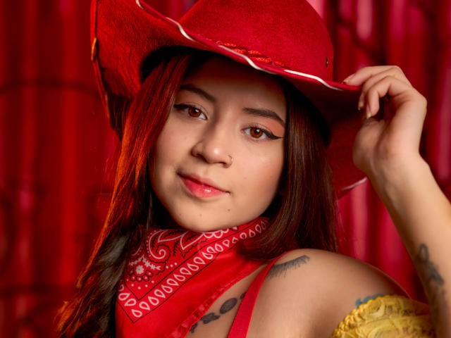 EmmaFeri with red cowboy hat, red bandana, and yellow off-shoulder top, has brown eyes, long brown hair, and tattoos, smiling slightly, holding hat with right hand.