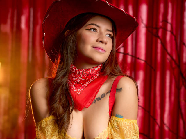EmmaFeri with brown hair, red bandana, yellow lace top, and red cowboy hat, showing her tits, tattoos on right shoulder, smiling, pink lipstick, red curtain background.