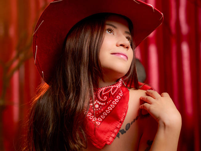 EmmaFeri with long brown hair, wearing a red cowboy hat and red bandana, looking up with a slight smile, hand near neck, showing a tattoo on her right side.