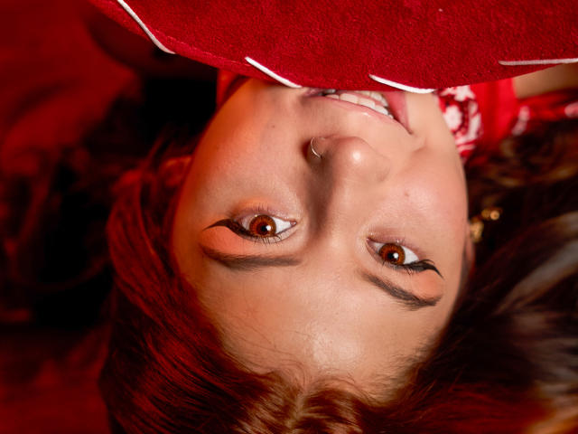 EmmaFeri's face, upside down, with red hair, brown eyes, black eyeliner, nose ring, and a smile, holding a red, textured fabric over her head. Her tits are partially visible.