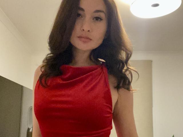 Dionisia, com cabelos castanhos ondulados, vestindo um vestido vermelho de veludo brilhante, mostrando os seios, olhando para a câmera com um leve sorriso em um ambiente bem iluminado..