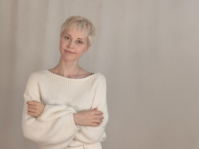 BarbaraRehfield with short blonde hair, smiling, wearing a white knitted sweater, arms crossed, standing against a plain background, looking cute and confident.